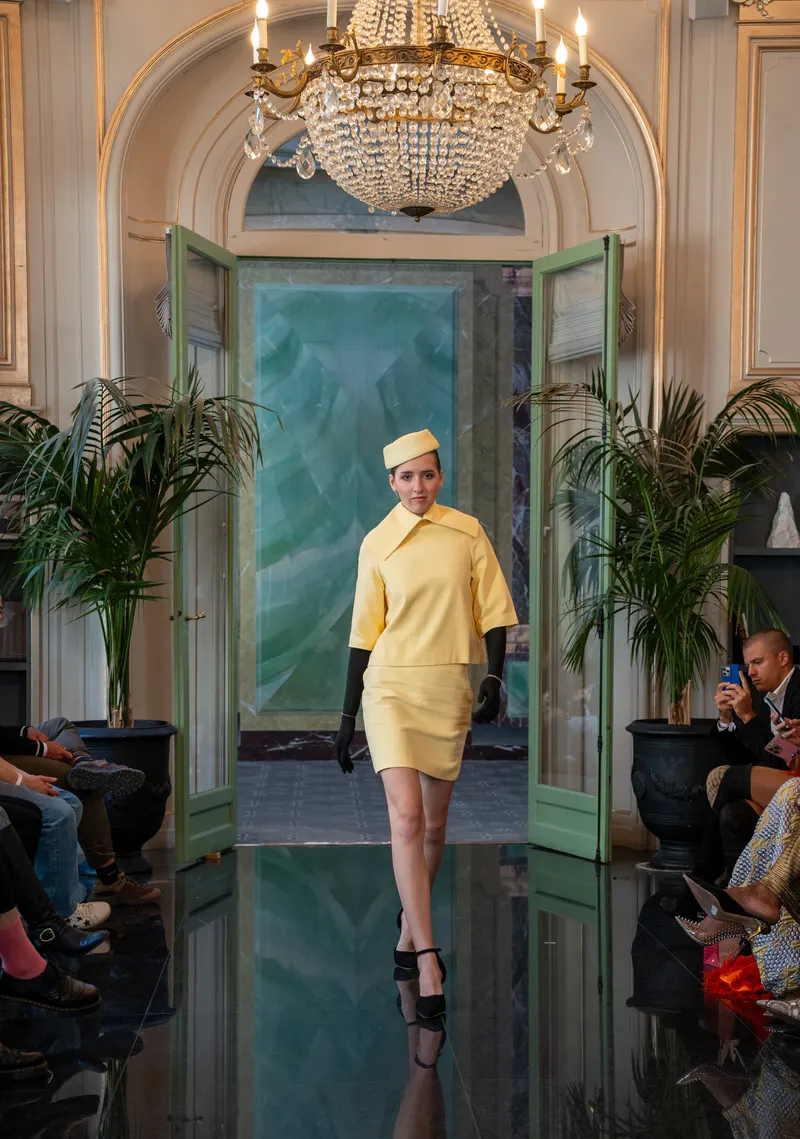 Lisa walking beneath a chandelier in a yellow suit, gloves and hat